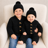 Baby and toddler on a cream chair in black Aaron crochet cardigans with buttons and pom-pom hats. Digital pattern PDF set.