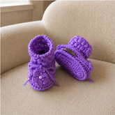 Purple Beth puff-stitch crochet booties on a cream chair, one standing and one lying down. Digital pattern PDF, Preemie–12M.