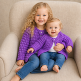 Girl and baby in blue jeans wear purple Beth puff-stitch crochet cardigans on a cream chair. Digital pattern PDF, NB–10Y.