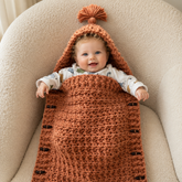Baby sitting in the Bodie rust chunky crochet hooded sleep sack with tassel and wooden toggles for a digital pattern PDF.