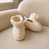 Cream Caleb cabled crochet baby booties on a cream chair, one standing and one lying down. Digital pattern PDF, Preemie–12M.