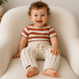 Baby in a rust and cream striped T-shirt wears cream Caleb cabled crochet pants on a cream chair. Digital pattern PDF, 0–2Y.