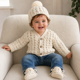 Baby in blue jeans on a cream chair wears cream Caleb crochet cardigan, hat and booties. Digital pattern PDF set, 0–2Y.