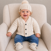 Baby in blue jeans on a cream chair wears the Carter crochet cardigan, hat and booties. Digital pattern PDF set, 0–2Y.