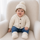Baby on a cream chair wears a cream Cooper cable-detail crochet cardigan, hat and booties. Digital pattern PDF set, 0–2Y.