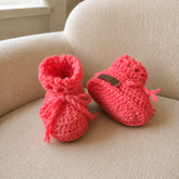Coral crochet baby booties on cream chair, one upright and one lying down to show the sole. Digital pattern PDF, Preemie–12M.