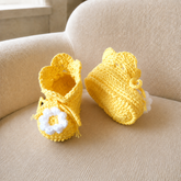 Yellow Daisy crochet baby booties on a cream chair, one standing and one lying down. Digital pattern PDF, Preemie–12M.