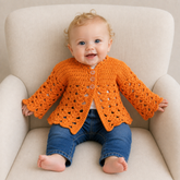 Baby wearing bright orange Dakota matinee crochet cardigan with blue jeans on a cream chair, size 0–3M, digital pattern PDF