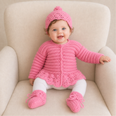 Baby wearing pink Emma peplum crochet cardigan, hat and booties with white tights on cream chair, 0–3M digital pattern PDF