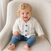 Baby on a cream chair wears white Ethan cabled V-neck crochet cardigan with blue tugboat buttons. Digital pattern PDF, 0–2Y.