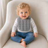 Baby in a T-shirt and jeans wears a silver Ethan cabled crochet bib on a cream chair. Digital pattern PDF.