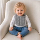 Baby in a T-shirt and jeans wears a silver Ethan cabled crochet bib on a cream chair. Digital pattern PDF.