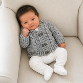 Newborn propped on a cream chair wears silver Ethan cabled V-neck crochet cardigan over a white onesie. Digital pattern PDF.