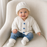 Baby in blue jeans on a cream chair wears white Ethan crochet cardigan, hat and booties. Digital pattern PDF set, 0–2Y.