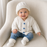 Baby in blue jeans on a cream chair wears white Ethan crochet cardigan, hat and booties. Digital pattern PDF set, 0–2Y.