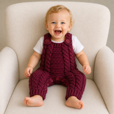 Laughing baby in a white T-shirt wears a maroon Hugo cabled crochet romper on a cream chair. Digital pattern PDF, NB–2Y.