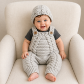Baby in a dark gray T-shirt wears a pale gray Hugo cabled crochet romper and hat on a cream chair. Digital pattern PDF set, 0–3M.
