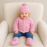 Baby wears pink Izzy crochet cardigan, pom-pom hat and booties with blue jeans on a cream chair; digital pattern PDF.