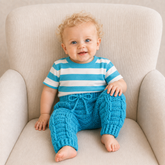Baby in a blue and white striped T-shirt wears blue Jack cabled crochet pants on a cream chair. Digital pattern PDF, NB–2Y.