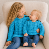 Older girl and baby in jeans wear blue Jack cabled crochet sweaters on a cream chair. Digital pattern PDF, Newborn–10Y.