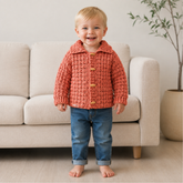 Happy toddler in coral James collared crochet cardigan with wood buttons, blue jeans by beige sofa, 0–2Y digital pattern PDF
