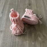 Pink Lola cabled crochet baby booties on wood, one upright and one lying down. Digital pattern PDF, Preemie–12M.