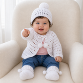 Baby pointing in white Lucy crochet cardigan, hat and booties with pink T-shirt and jeans on cream chair, 0–3M digital pattern PDF