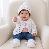 Baby pointing in white Lucy crochet cardigan, hat and booties with pink T-shirt and jeans on cream chair, 0–3M digital pattern PDF