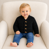 Baby wearing black Aaron V-neck crochet cardigan with blue jeans on a cream chair, sizes 0–3Y, digital pattern PDF