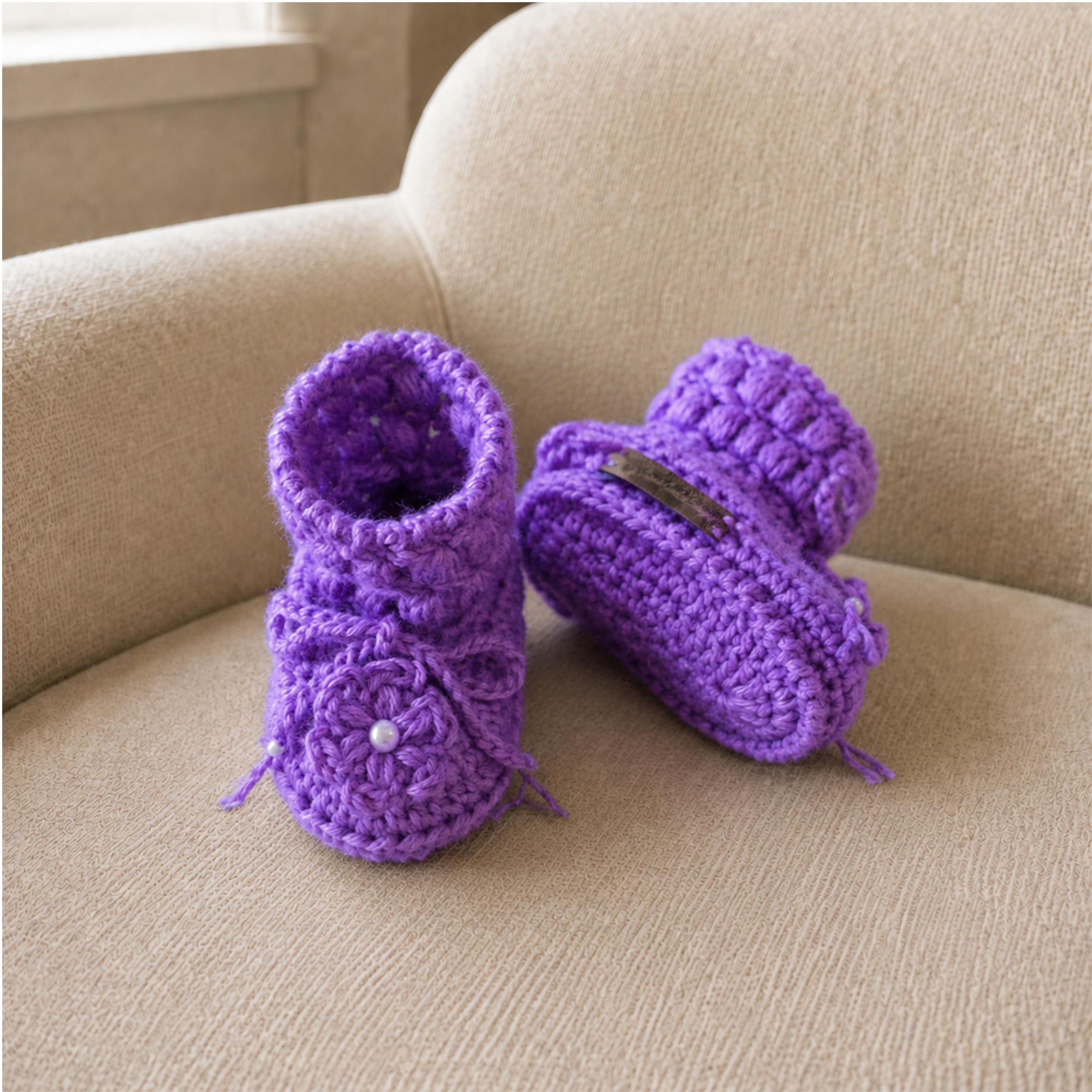 Purple Beth puff-stitch crochet booties on a cream chair, one standing and one lying down. Digital pattern PDF, Preemie–12M.