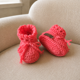 Coral crochet baby booties on cream chair, one upright and one lying down to show the sole. Digital pattern PDF, Preemie–12M.
