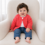 Baby in a white T-shirt and jeans wears the coral-red Coral crochet cardigan on a cream chair. Digital pattern PDF, 0–2Y.