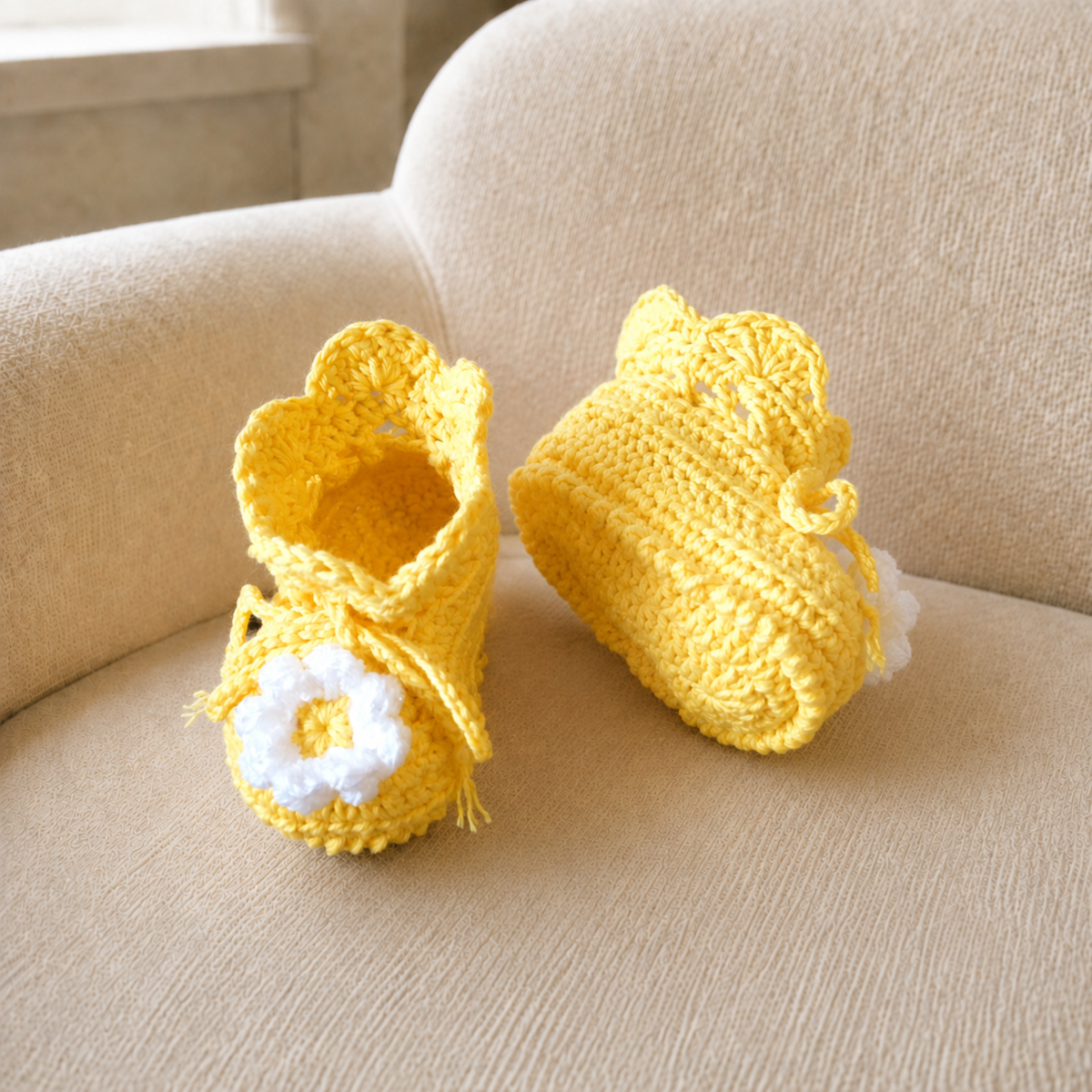 Yellow Daisy crochet baby booties on a cream chair, one standing and one lying down. Digital pattern PDF, Preemie–12M.