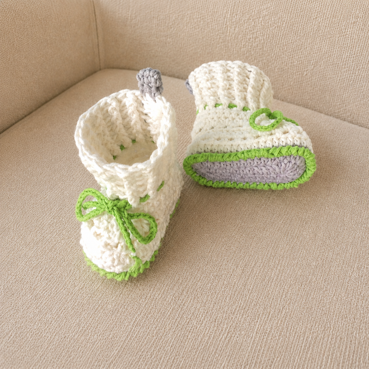 Ivy white, gray and green cuffed crochet booties on cream chair, one upright and one lying down. Digital pattern PDF, NB–12M.