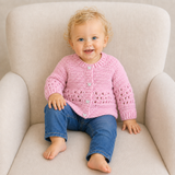 Baby wears pink Izzy crochet cardigan with blue jeans on a cream chair; digital pattern PDF, 0–2Y.