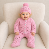 Baby wears pink Izzy crochet cardigan, hat, booties and pants on a cream chair; digital pattern PDF, 0–2Y.