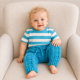 Baby in a blue and white striped T-shirt wears blue Jack cabled crochet pants on a cream chair. Digital pattern PDF, NB–2Y.