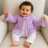 Baby in a floral outfit wears a mauve Orchid tie-front crochet cardigan on a cream chair. Digital pattern PDF, NB–12M.