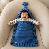 Baby lying in the River blue cabled crochet cocoon with arms out and wooden side toggles for a digital pattern PDF.
