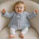 Baby in white pants wears a Sky tie-front crochet matinee jacket on a cream chair. Digital pattern PDF, 0–3M.